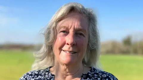 Stuart Woodward/BBC Jackie Thacker wearing a black dress with white flowers on it. She has grey hair and is smiling while standing in a field.
