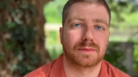 Emma Baugh/BBC Paddy wearing an orange button-down shirt, looking at the camera. He appears to be sitting outside, with greenery behind him.