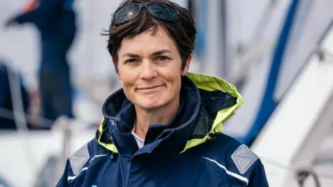 EMCTrust Dame Ellen MacArthur wearing a blue sailing jacket looking straight at the camera. She has short brown hair and sunglasses on top of her head.