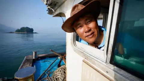 BBC/Hosu Lee A man wearing a hat sticks his head out of the window of a boat. In the background is sea and an island.